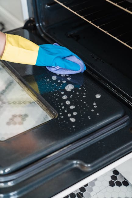 Close-up of a person's hand wearing a blue cleaning glove and a yellow apron, wiping the black glass surface of an oven door with a blue cloth. The oven's interior rack is visible, and the surface sho