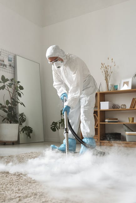 A professional cleaner from Cleaners Richmond upon Thames is performing surface cleaning in a residential living room. The cleaner is wearing a full white protective suit, face mask, and blue gloves, and is operating a steam or fogging device that emits white mist onto the beige carpeted floor. The room features a wooden bookshelf with various decorative items, a mirror leaning against the wall, and potted plants, with soft natural lighting illuminating the space. The scene emphasizes thorough sanitisation or deep cleaning of carpets and surfaces within a clean, well-organized indoor environment. This image illustrates the use of specialised equipment for maintaining hygiene and cleanliness as part of domestic cleaning services provided by [COMPANY_NAME].
