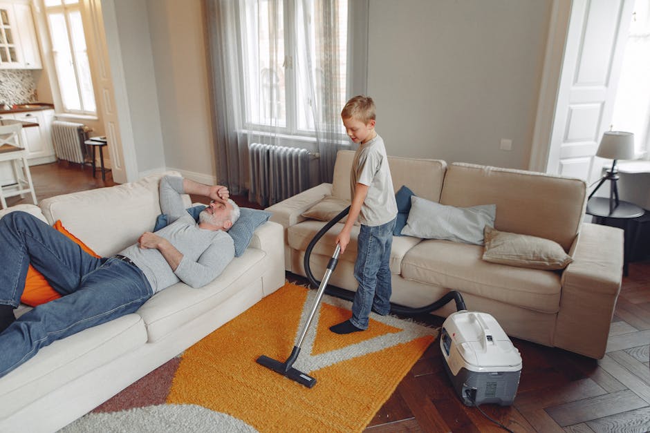 A professional cleaner from Cleaners Richmond upon Thames is performing surface cleaning in a residential living room. The cleaner is wearing a full white protective suit, face mask, and blue gloves, and is operating a steam or fogging device that emits white mist onto the beige carpeted floor. The room features a wooden bookshelf with various decorative items, a mirror leaning against the wall, and potted plants, with soft natural lighting illuminating the space. The scene emphasizes thorough sanitisation or deep cleaning of carpets and surfaces within a clean, well-organized indoor environment. This image illustrates the use of specialised equipment for maintaining hygiene and cleanliness as part of domestic cleaning services provided by [COMPANY_NAME].