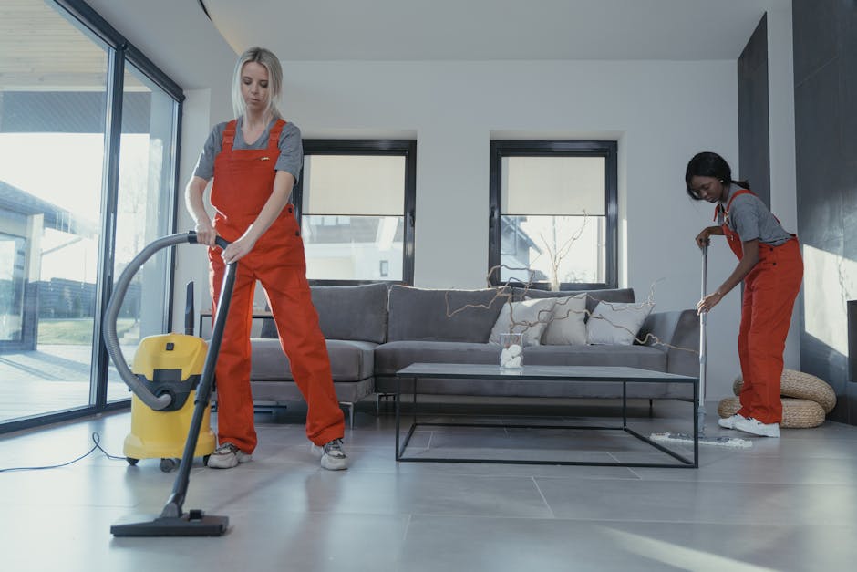 Two professional cleaners from Cleaners Richmond upon Thames wearing gray t-shirts and orange overalls perform surface cleaning in a modern, well-lit living room. The cleaner on the left uses a yellow vacuum cleaner to clean a dark gray tiled floor, while the cleaner on the right mops a light-colored, textured area rug near a gray fabric sofa with decorative cushions. The room features large glass sliding doors, allowing natural light to illuminate the space, with a coffee table and minimalist decor visible. The surfaces, including the tiled floor and fabric upholstery, appear clean and well-maintained, reflecting thorough domestic cleaning efforts aligned with the Richmond Hill end of tenancy cleaning guide for landlords.